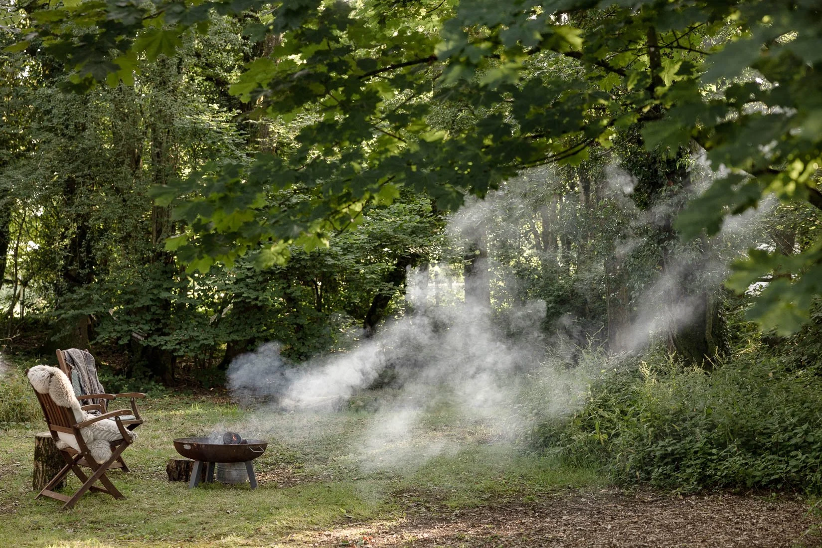 A peaceful outdoor scene with two wooden chairs draped with cozy blankets near a small fire pit, surrounded by lush green trees and bushes, with smoke rising into the air.
