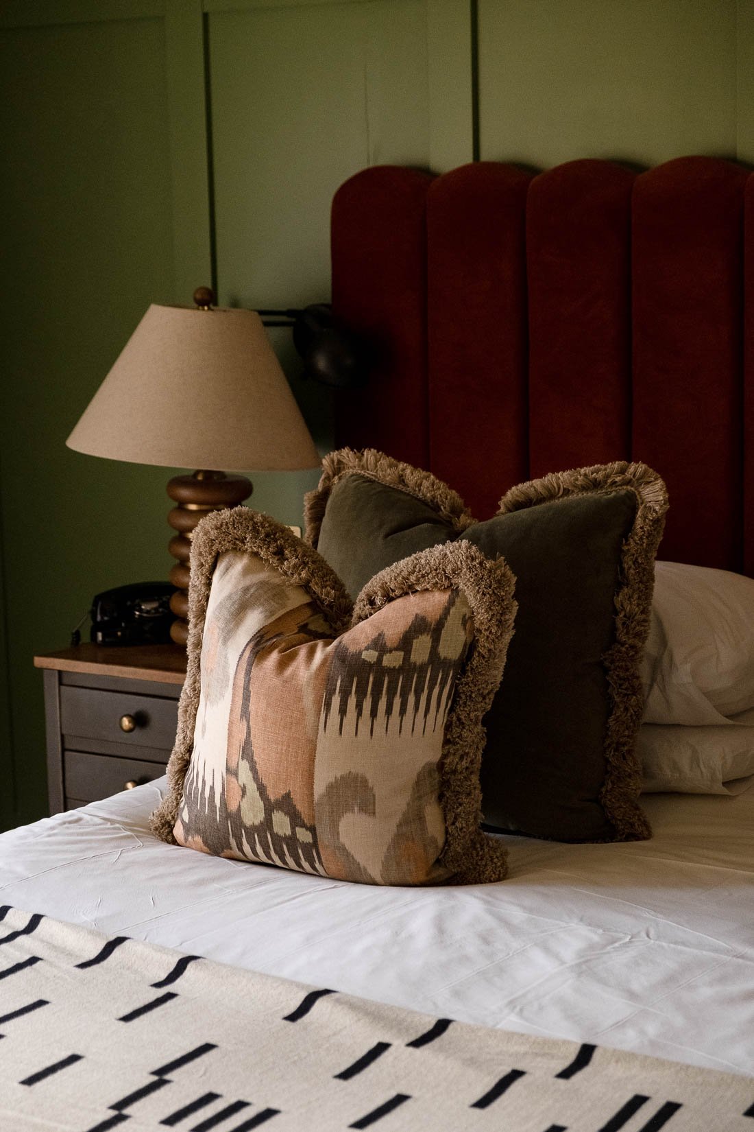A bedroom with a white bedspread, decorative pillows with fur edges, a wooden nightstand, a beige table lamp, and a green wall with a red upholstered headboard.