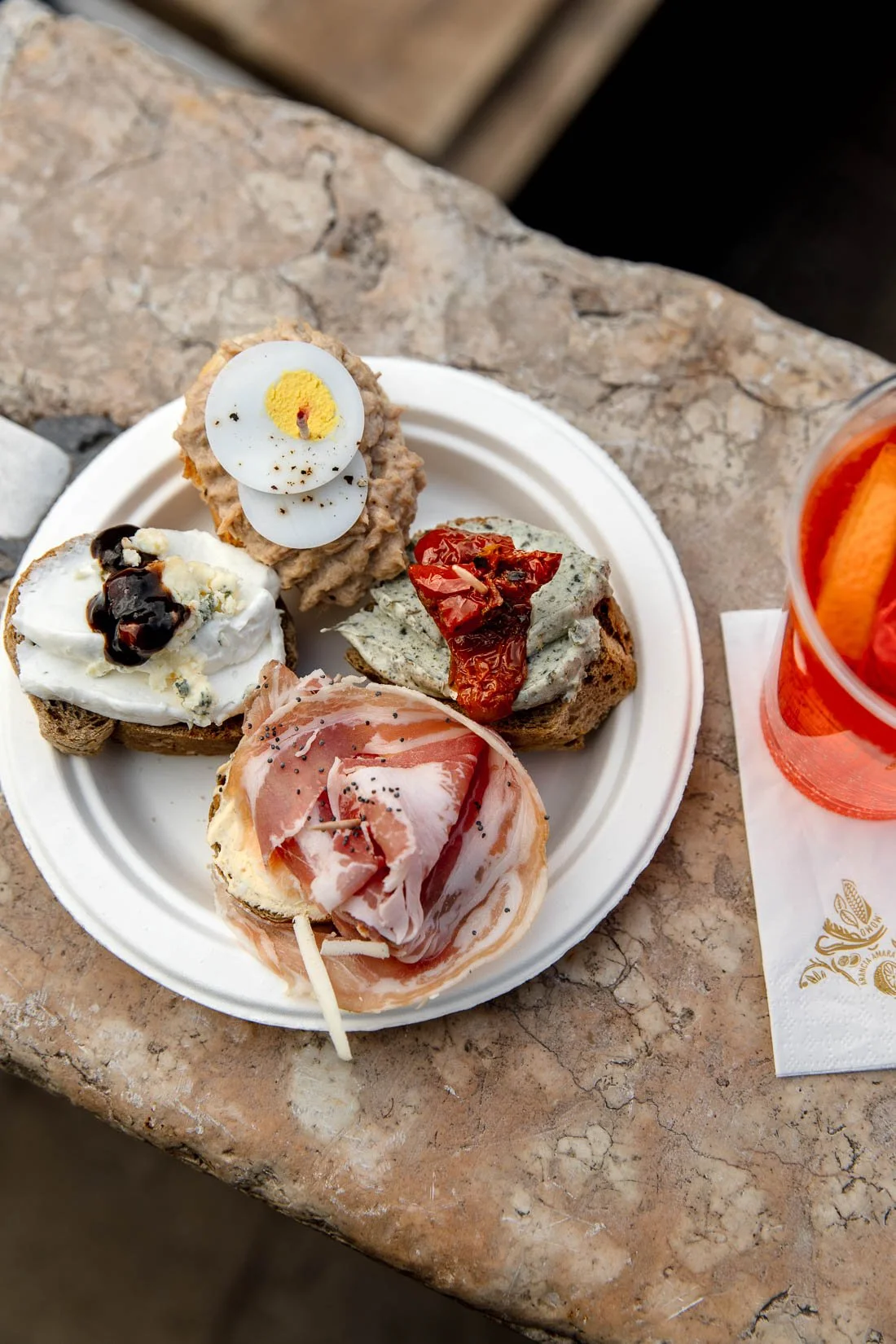 Assorted tapas on a white plate, including bread topped with various spreads, cheese, cured ham, and eggs, with a glass of orange beverage on the side.