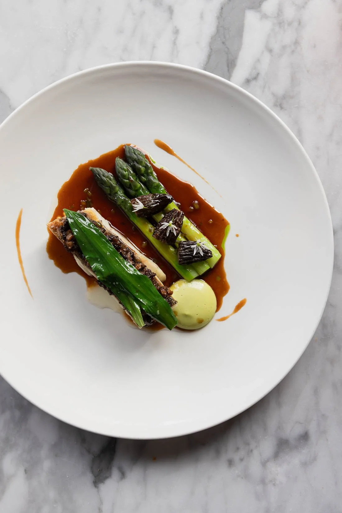Looking down at a plated gourmet dish with asparagus, a green vegetable, and a piece of fish in a brown sauce with a dollop of green puree, garnished with edible flowers, on a white plate on a marble surface.