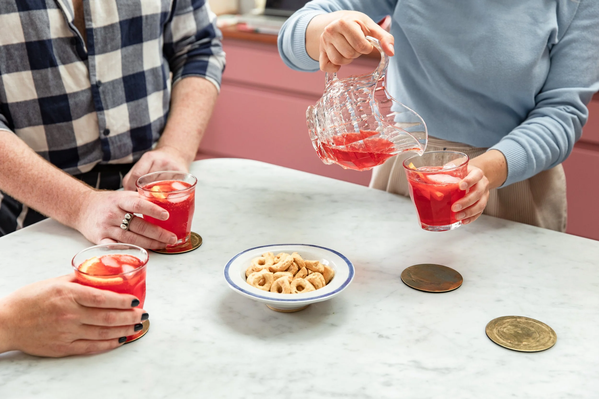 Two people are serving pink punch from a glass pitcher into clear glasses with ice and lemon slices on a white marble table, with a bowl of pretzels and gold coins in the scene.
