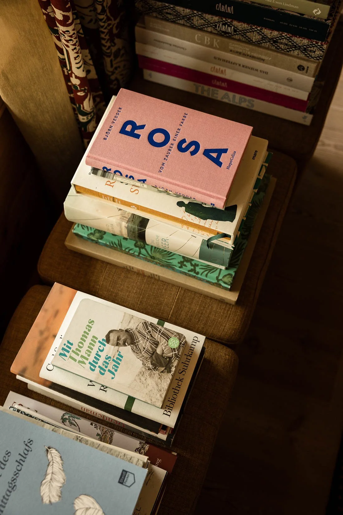 A stack of books on a wooden chair and nearby surface, with additional books on a shelf in the background. The top book on the chair is pink with blue title text.