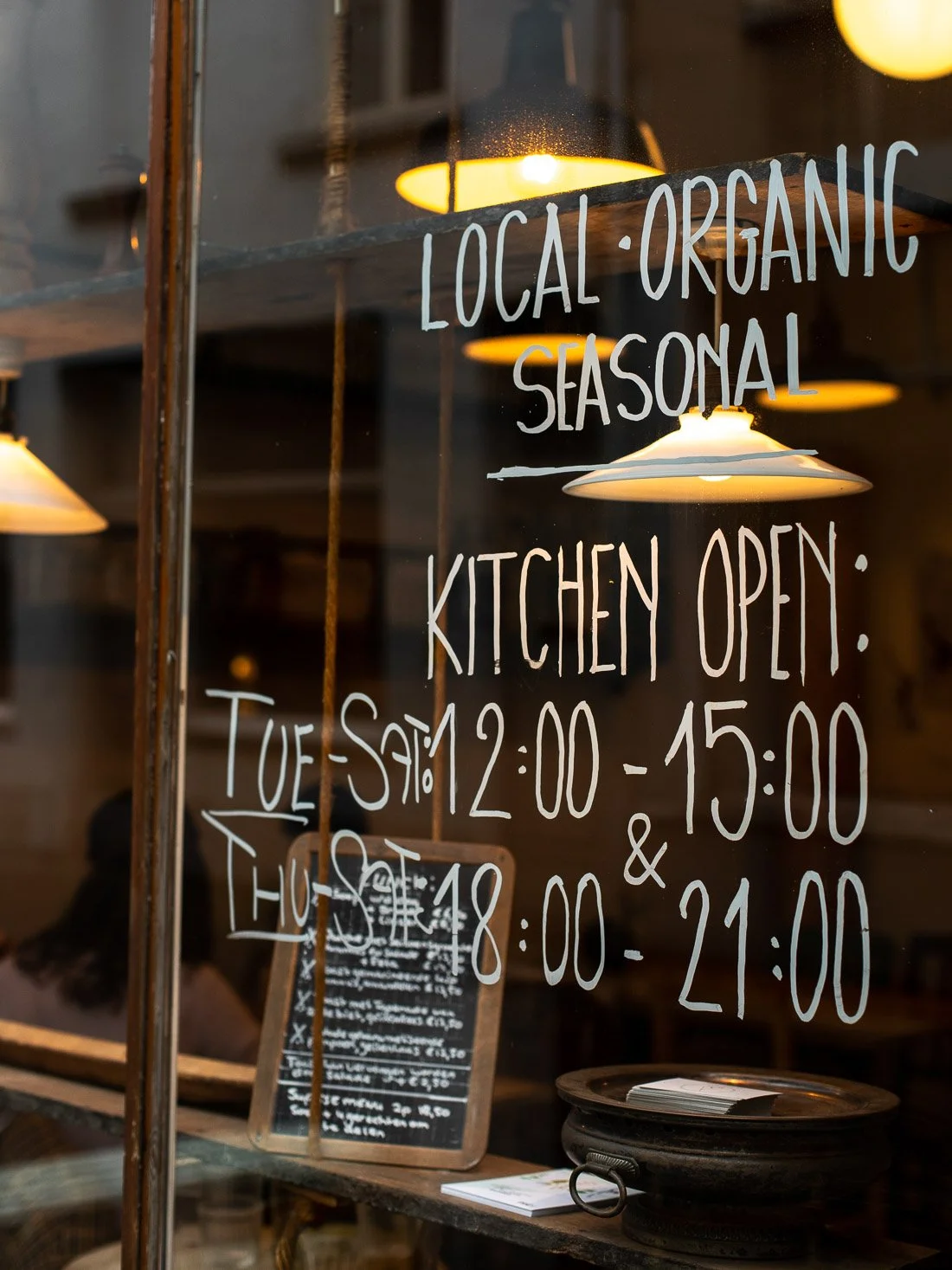 A glass window with handwritten sign in white lettering indicating the shop offers local, organic, seasonal products. The shop's live hours are Tuesday to Saturday from 12:00 PM to 3:00 PM and Thursday to Friday from 6:00 PM to 9:00 PM. Inside, warm 