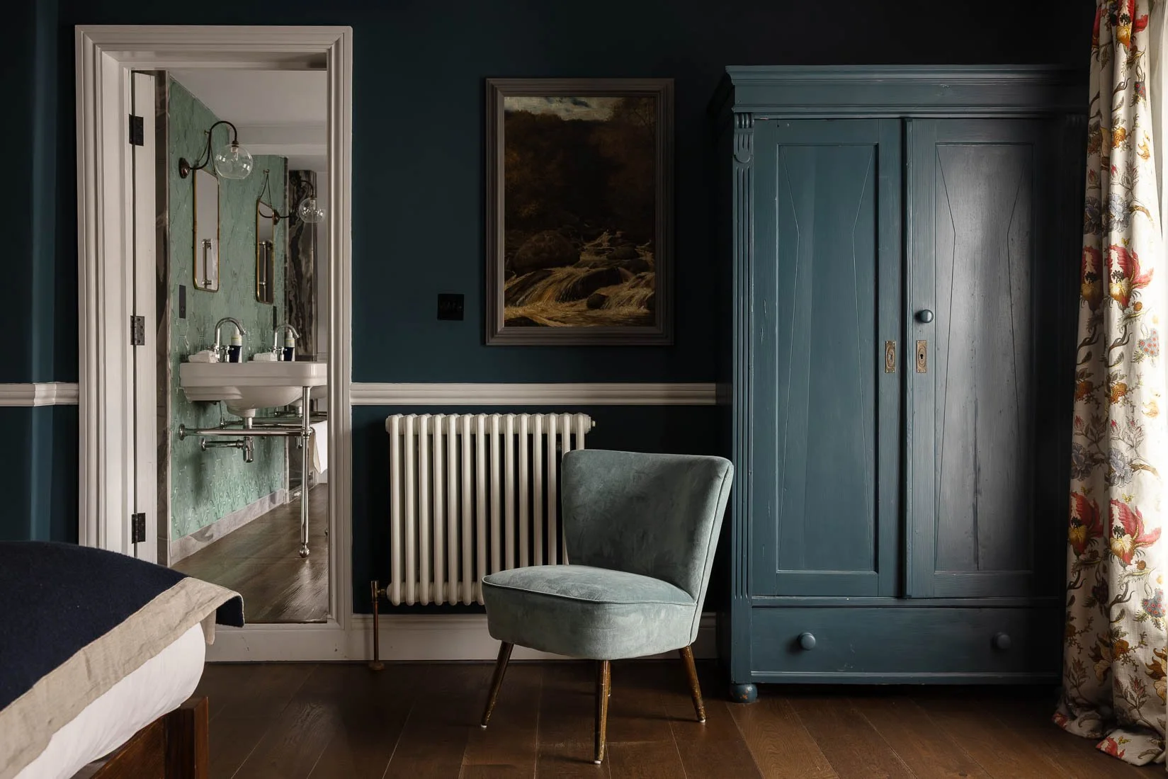 Interior view of a room showing a blue cabinet, an armchair, a radiator, a painting, and a doorway to a bathroom with green wallpaper and double sinks.