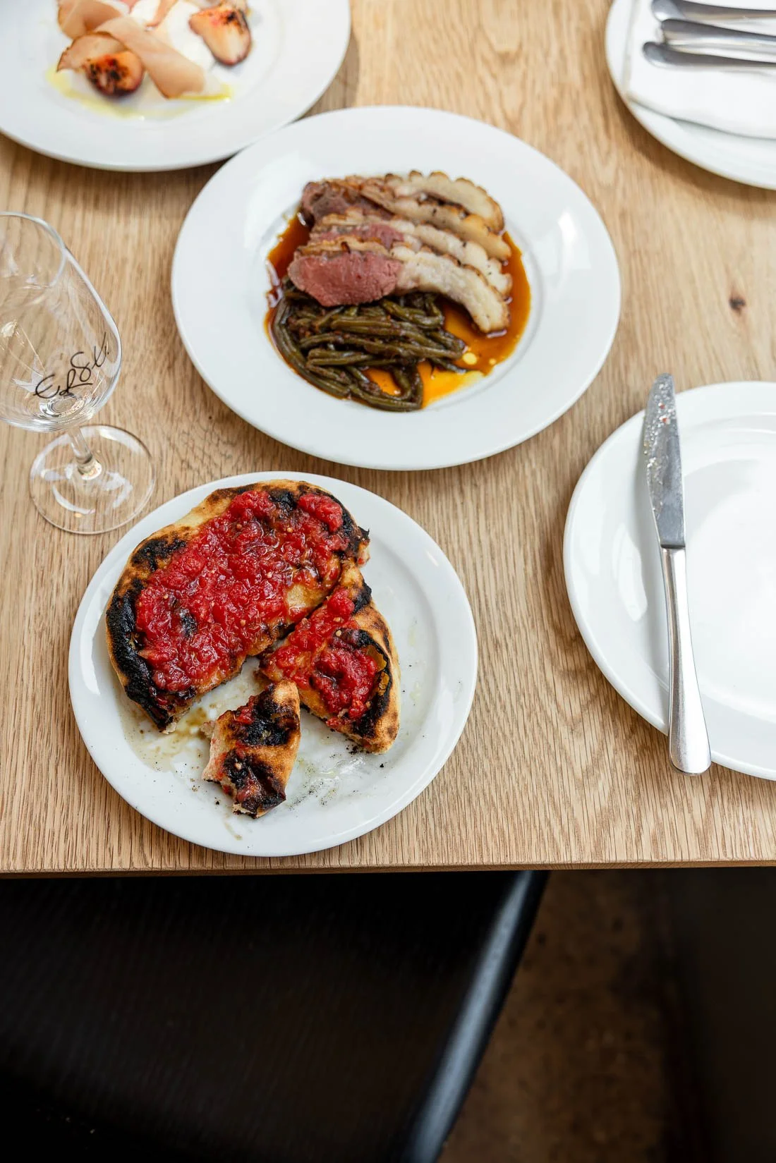 A table with Italian dishes, including a partially eaten pizza with tomato sauce, a plate with sliced steak topped with cooked vegetables, and a plate with what appears to be grilled meat wrapped around a filling, set on white plates with silverware 