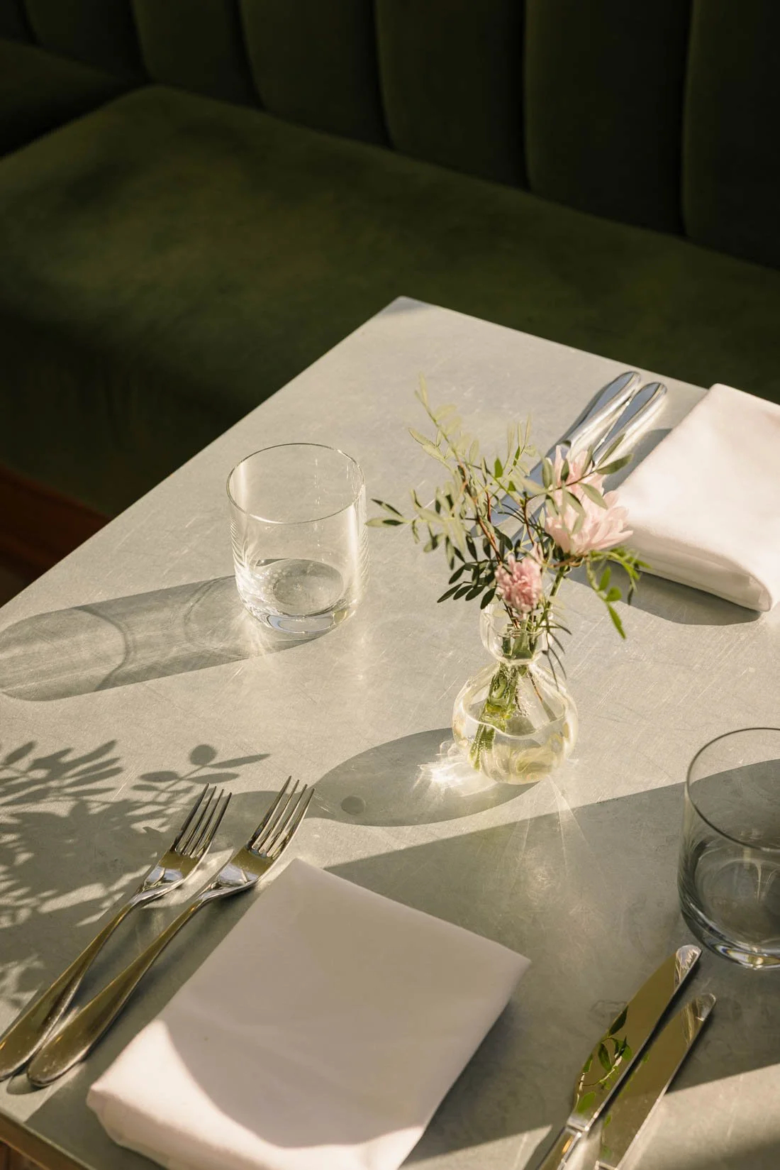 Elegant dining table set with a white tablecloth, two forks, a knife, a glass of water, a folded white napkin, and a small vase with pink flowers and green foliage. Sunlight casts shadows on the table.