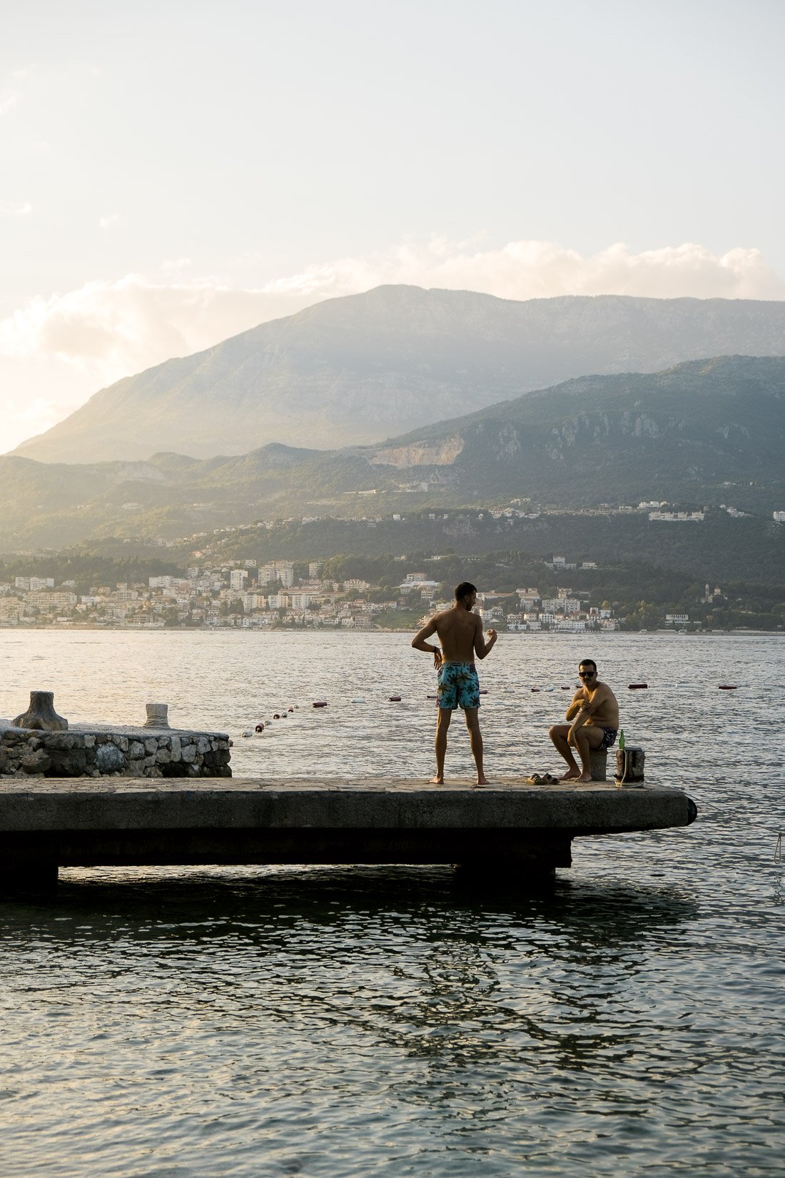 Two men on a dock by the water, one standing and talking, the other sitting with a bottle beside him, with mountains and a town in the background.
