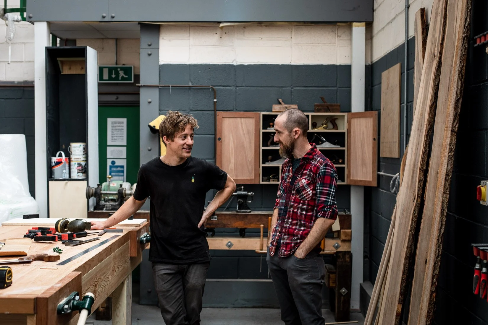 Two men talking in a woodworking shop. The man on the left has curly hair and is wearing a black t-shirt, the man on the right has a beard and is wearing a red plaid shirt. The shop has wood planks on the right and a workbench with tools and supplies