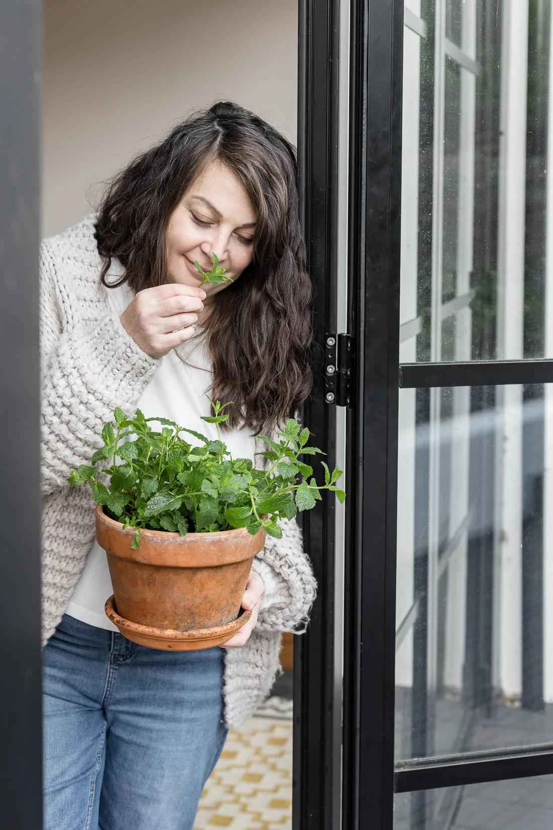 A woman with long curly hair standing by a glass door, holding a terracotta pot with green mint plants. She is smelling a mint leaf.