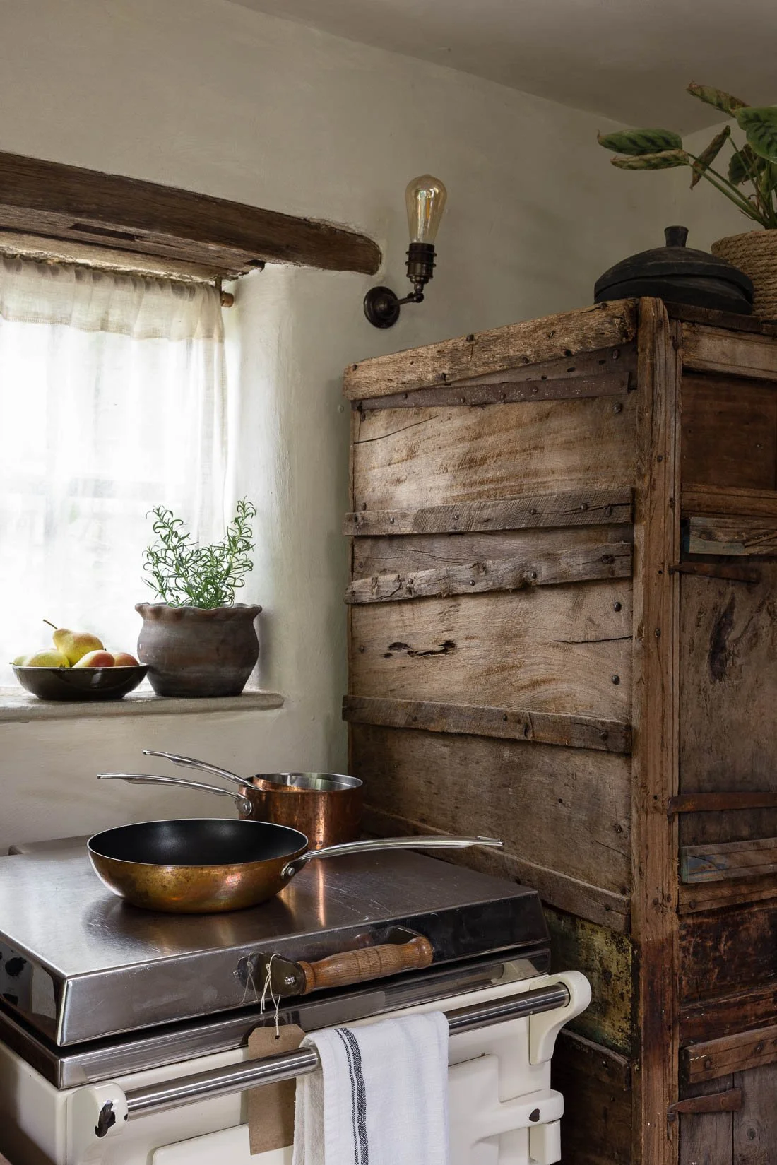 Rustic kitchen corner with a window, a potted plant, a bowl of pears, a vintage wooden cabinet, a wall-mounted light bulb, a stainless steel stove with a frying pan and copper pot on top, and a white towel hanging from the stove.