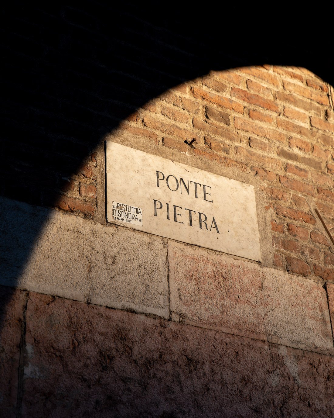 Street sign on a brick wall that reads 'Ponte Pietra' with a smaller sticker in Italian.