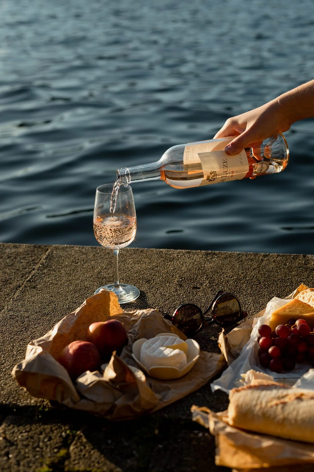 A hand pouring rosé wine into a glass near a body of water, with picnic items including grapes, cheese, bread, sunglasses, and a pastry on the ground.