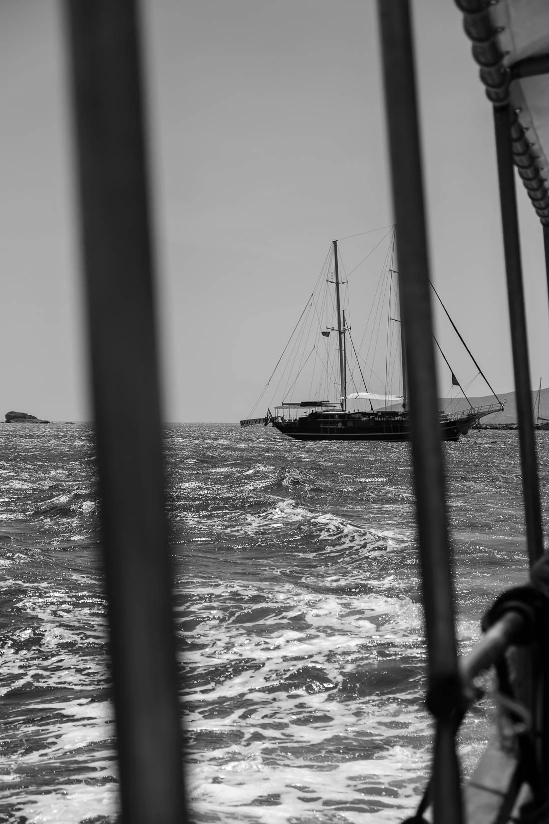 Black and white photo taken from a boat showing a large sailing yacht on the water, framed by parts of the boat's railings.