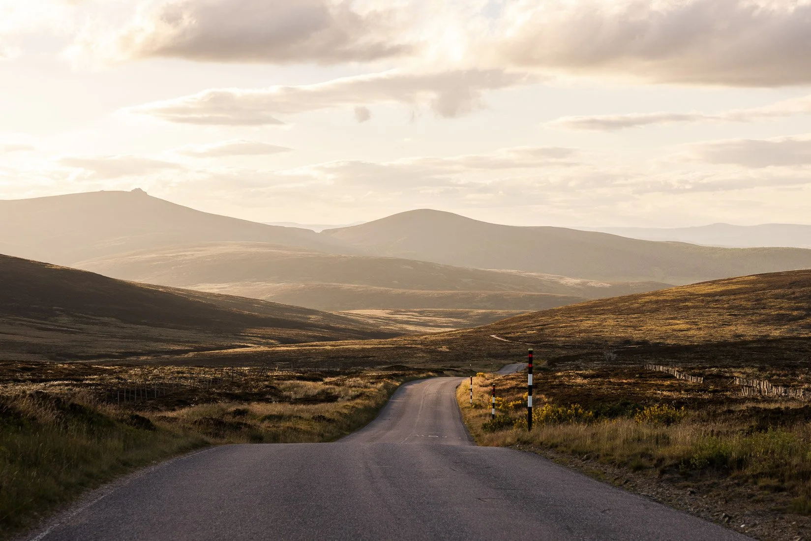 A winding rural road cutting through a hilly landscape with grassy fields and distant mountains under a cloudy sky.