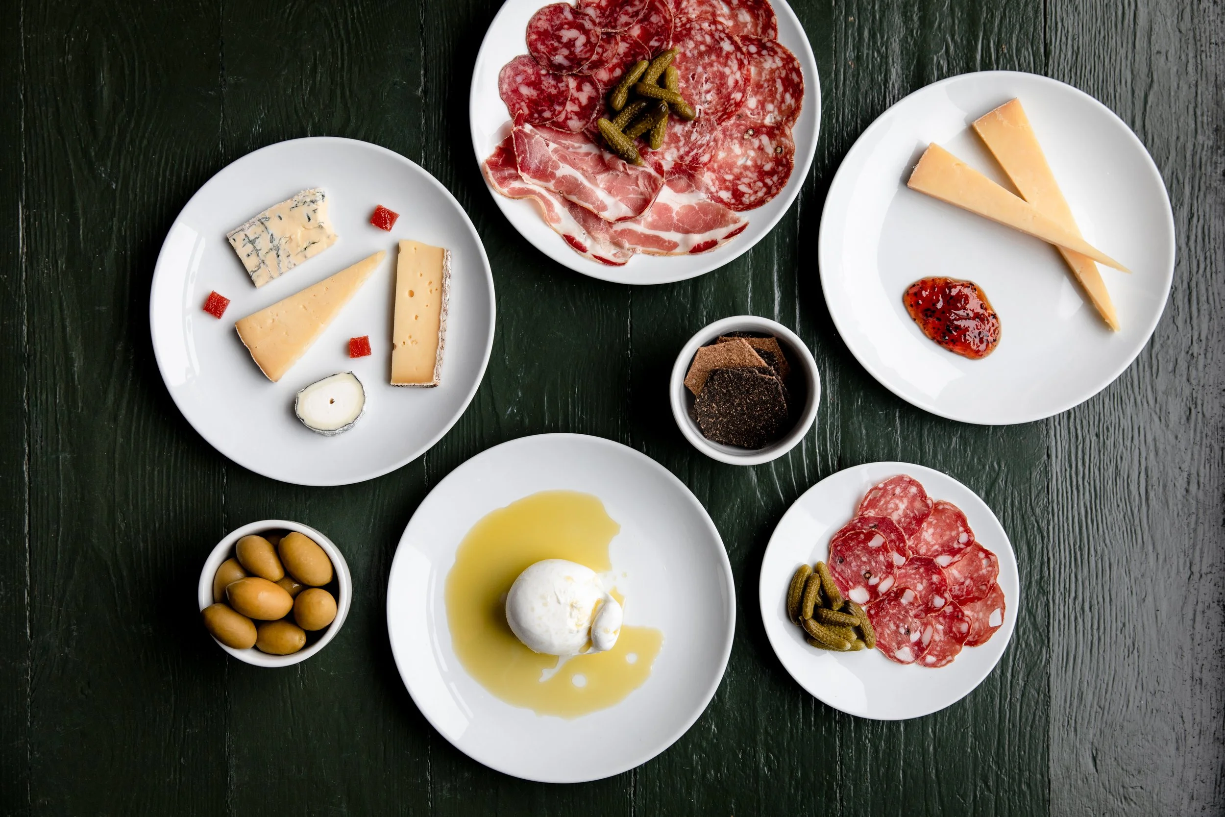 Various cheese, cured meats, olives, and condiments arranged on white plates on a dark green wooden surface.