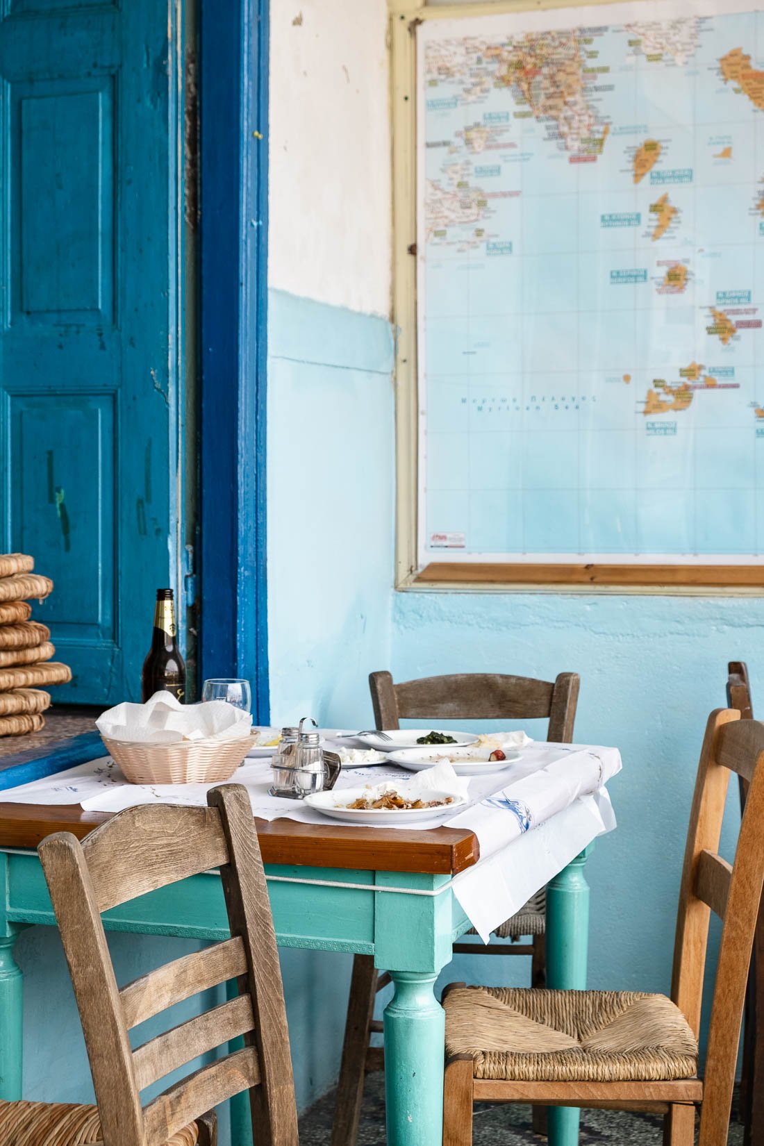 A cozy restaurant interior with a light blue wall, a map, a blue door, and a wooden table with chairs. The table has used plates with leftover food, utensils, a basket with napkins, a glass, a bottle, and salt and pepper shakers.