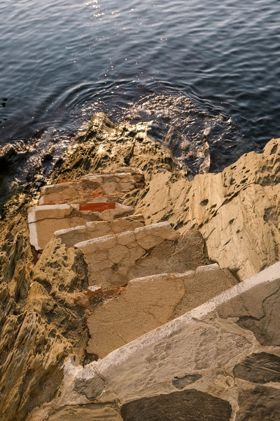 Close-up of rocky shoreline with beige and brown stones leading to water with small waves and reflections.