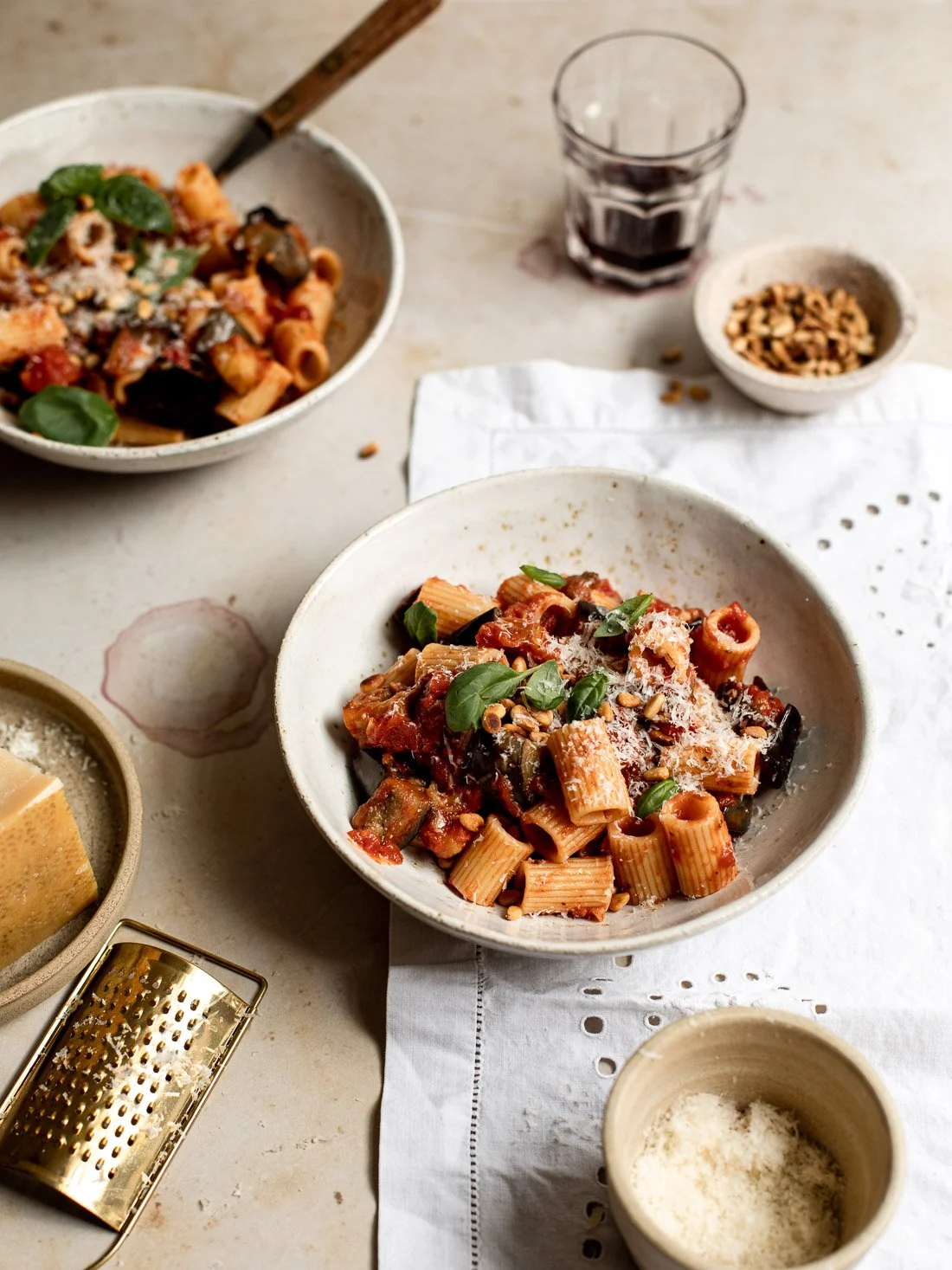 Two bowls of pasta with tomato sauce, topped with basil, grated cheese, and pine nuts, on a white table setting with a glass of red wine, small bowls, and a cheese grater.