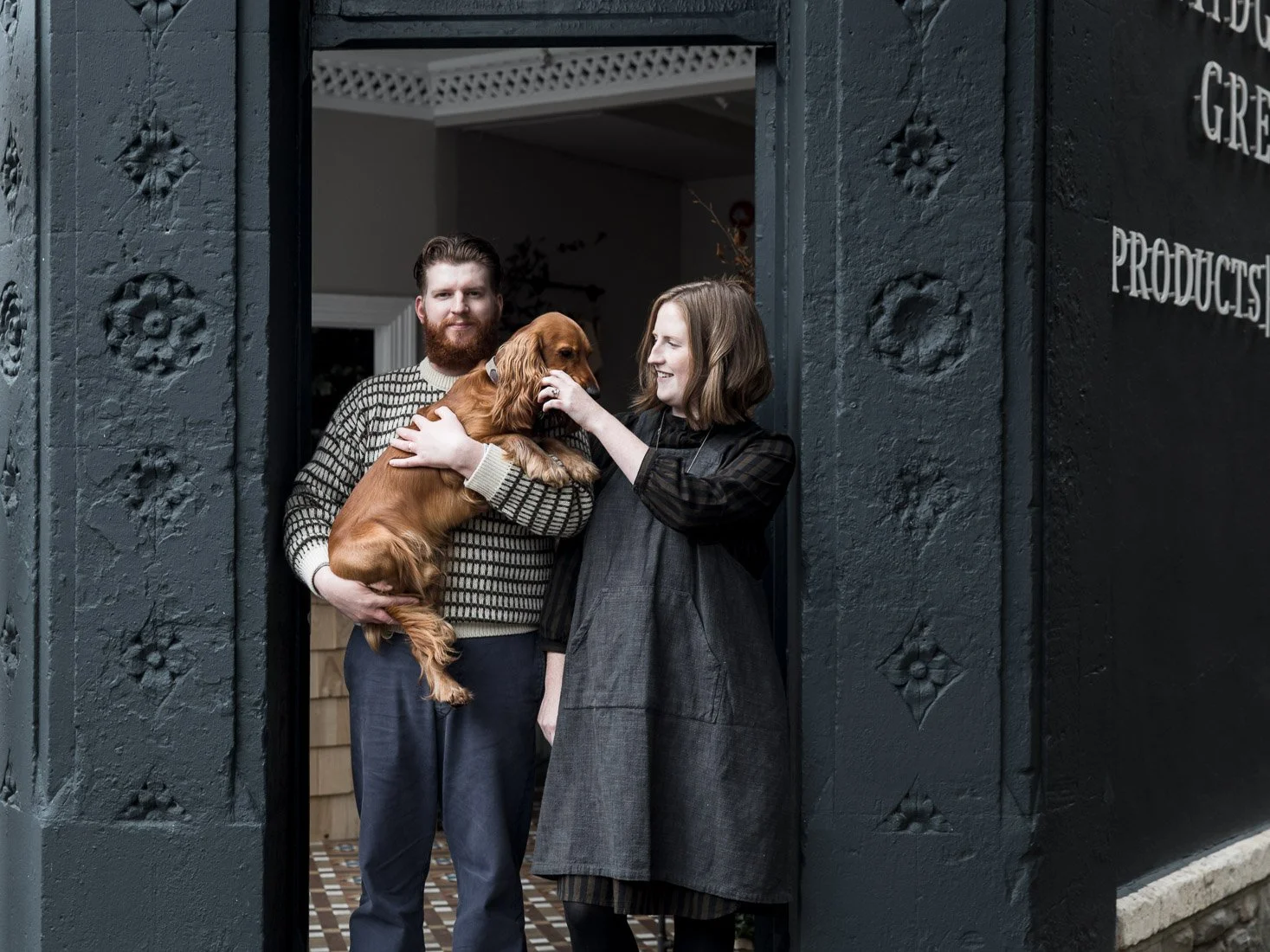 A man and woman stand at the doorway of a dark building, holding and petting a brown dog between them. The man has a beard and is wearing a patterned sweater, while the woman has shoulder-length hair and is dressed in a dark dress and apron. The buil