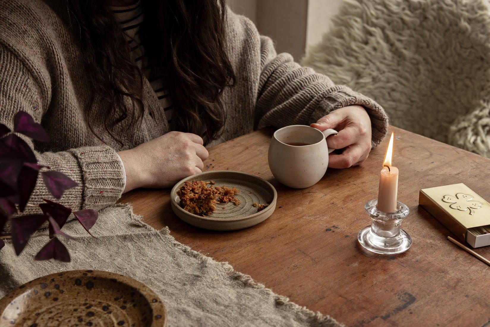 A woman sitting at a wooden table with a beige sweater, holding a mug, with a plate of food, a lit candle, and a matchbox nearby.