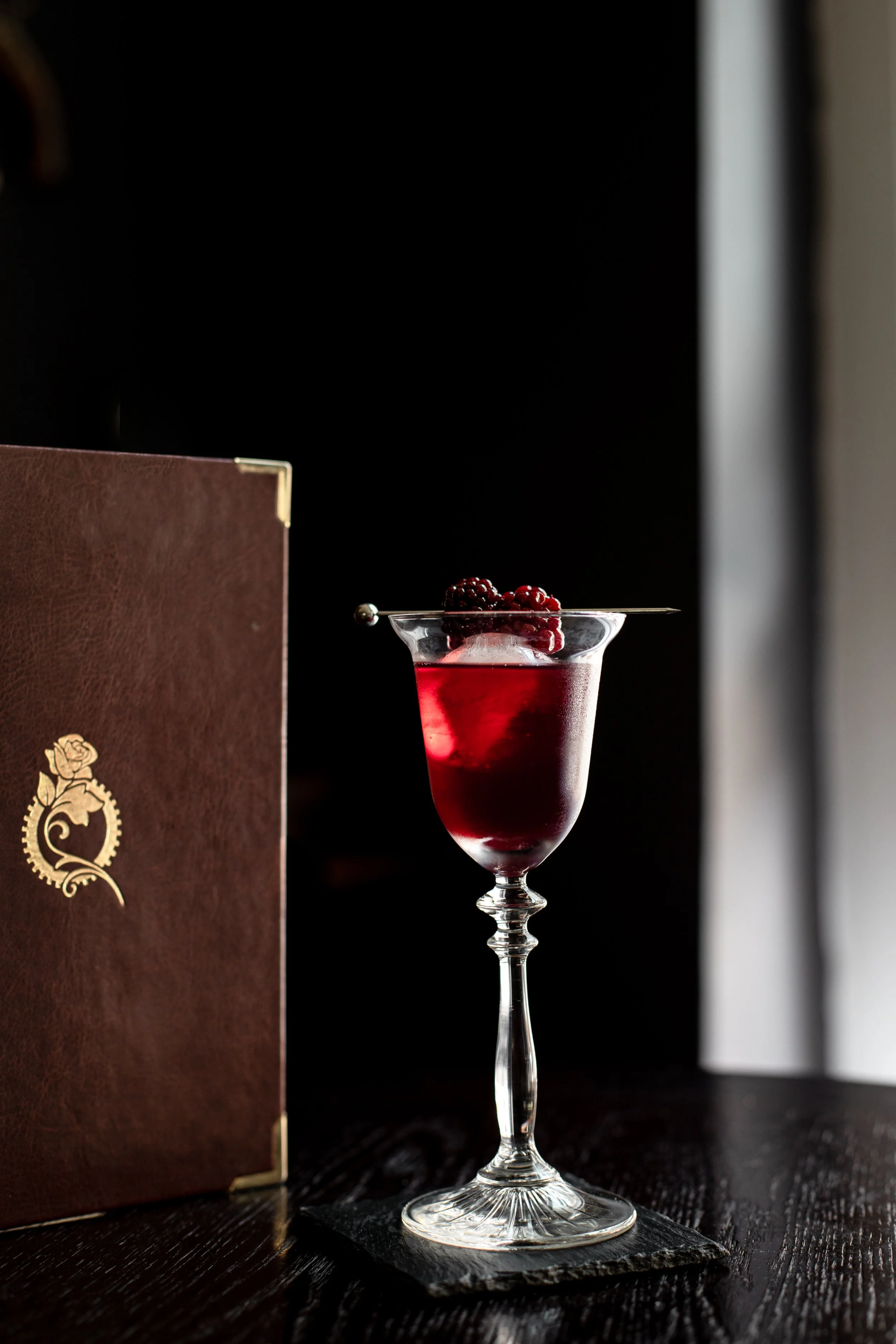 A tall, elegant glass filled with red berry cocktail garnished with blackberries on top, set on a black slate coaster on a dark wooden table, with a brown menu on the left and a black and gray background.