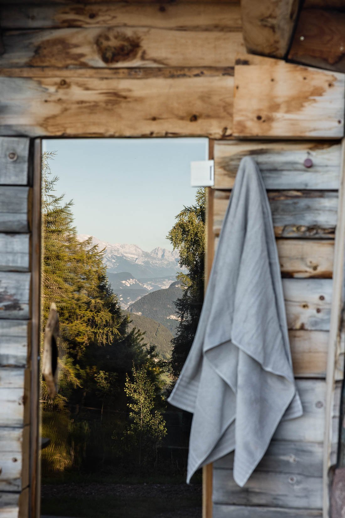 View of mountains and trees seen through a small window in a wooden cabin, with a towel hanging on the side.