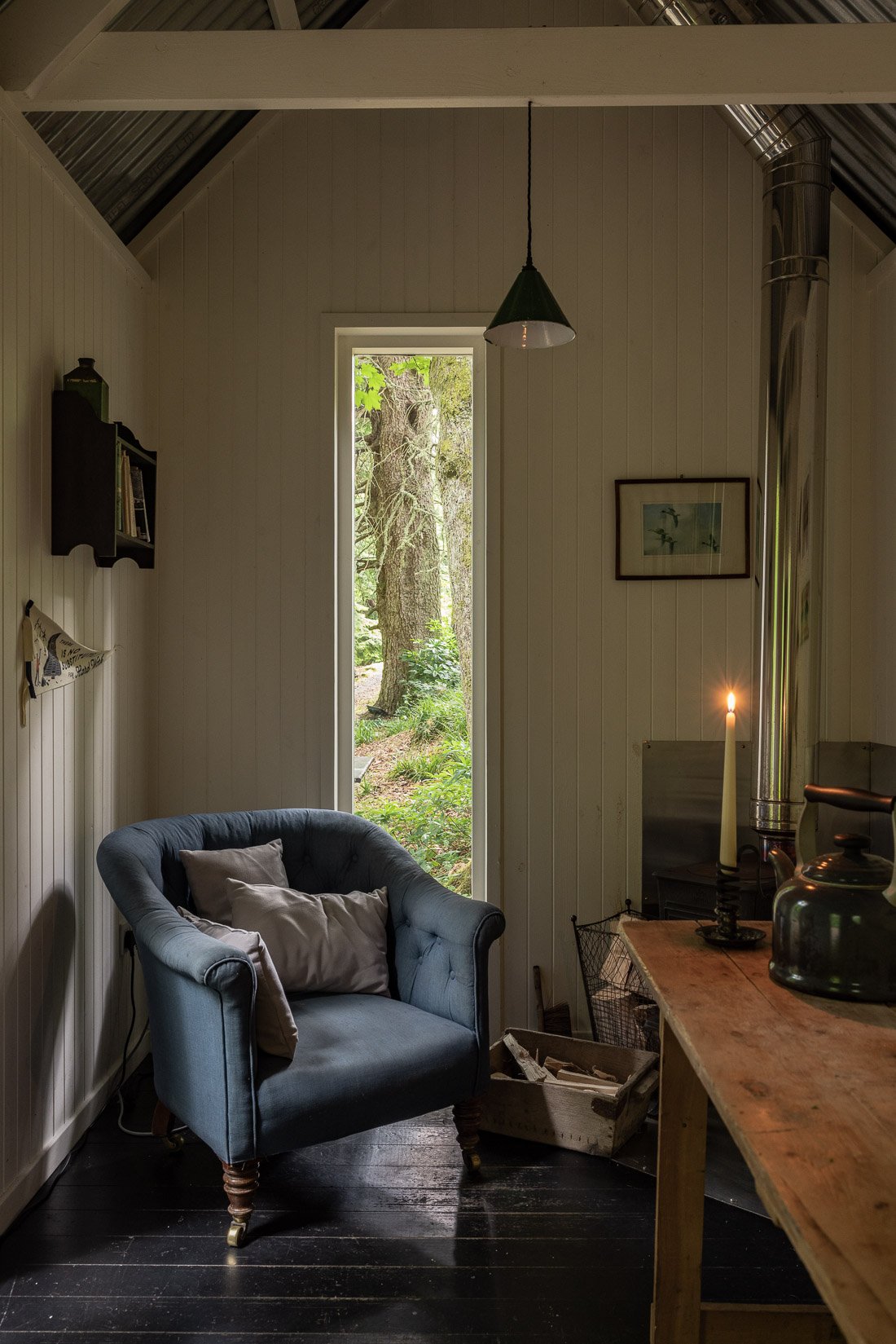 A cozy indoor space with a blue armchair, light-colored pillows, a candle on a black candle holder, a wooden table, a small bookshelf on the wall, and a large window showing trees and greenery outside.