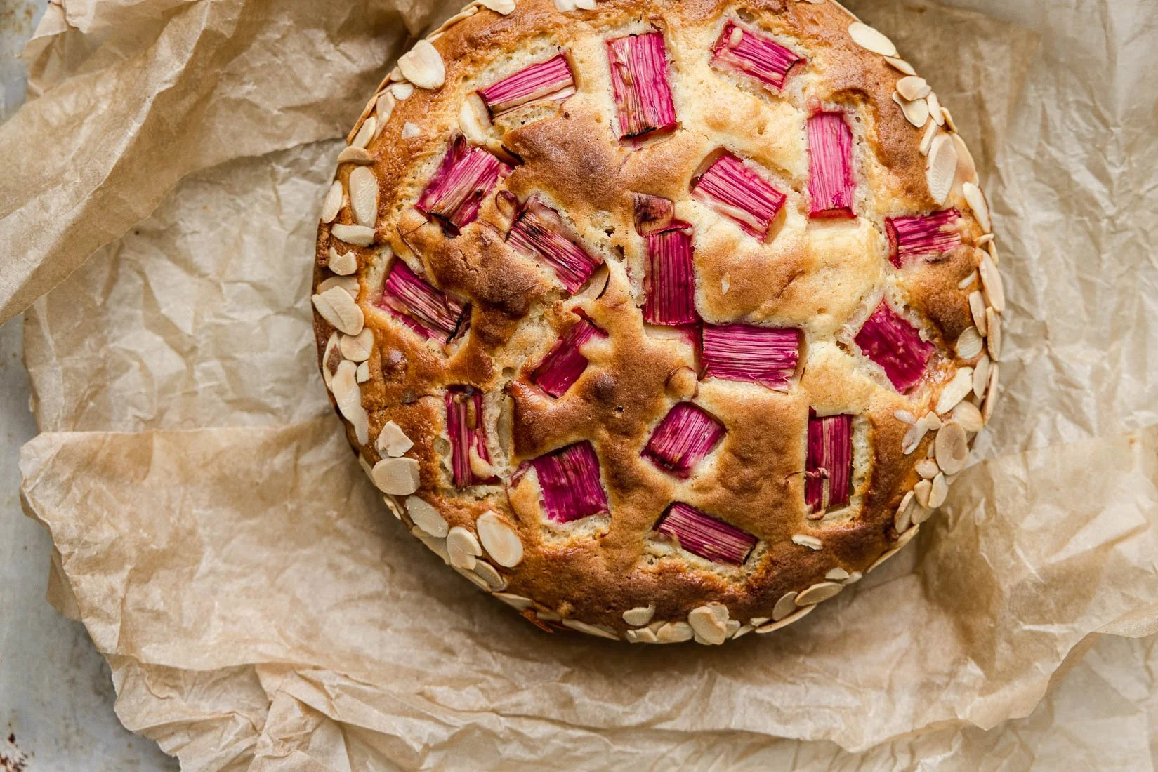 A round cake with pink rhubarb chunks and sliced almonds on top, resting on crumpled parchment paper.