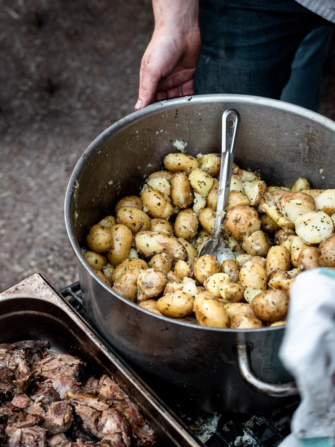 A person is holding a large metal pot filled with small roasted potatoes with herbs, with a serving spoon inside. The pot is on an outdoor grill next to some cooked meat.