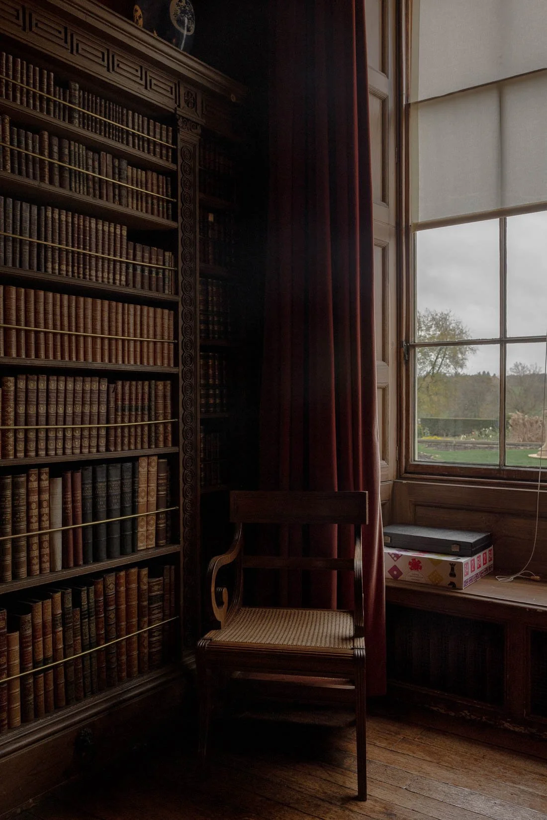 Interior of a room with tall wooden bookshelves filled with leather-bound books, an empty wooden chair with a woven seat, a large window with red curtains, and a view of trees and sky outside.