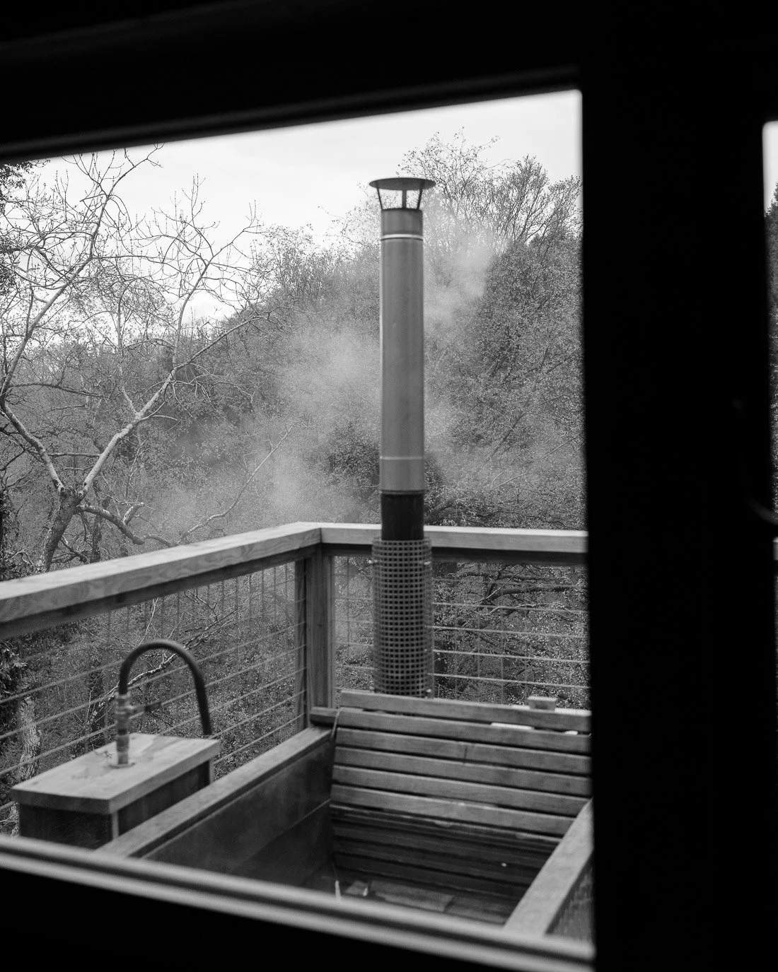 View of a wooden balcony with a bench, and a wood-fired outdoor heater, seen through a window, with trees in the background.