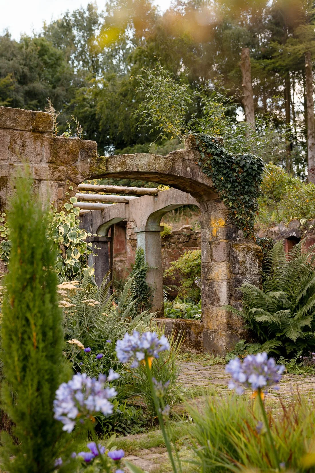 An old stone and brick garden structure with arches, surrounded by lush green plants and purple flowers, in a peaceful outdoor setting.