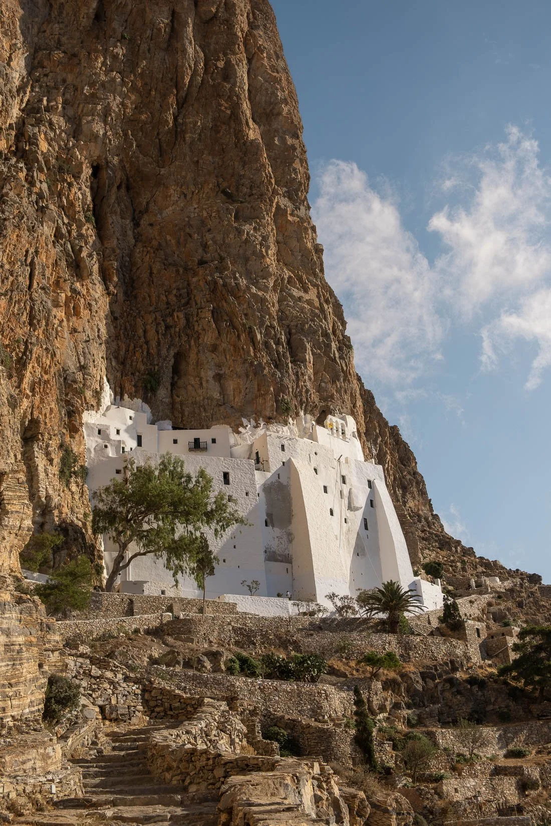 White buildings built into a rocky cliffside, with stone pathways and stairs leading up, under a clear blue sky.