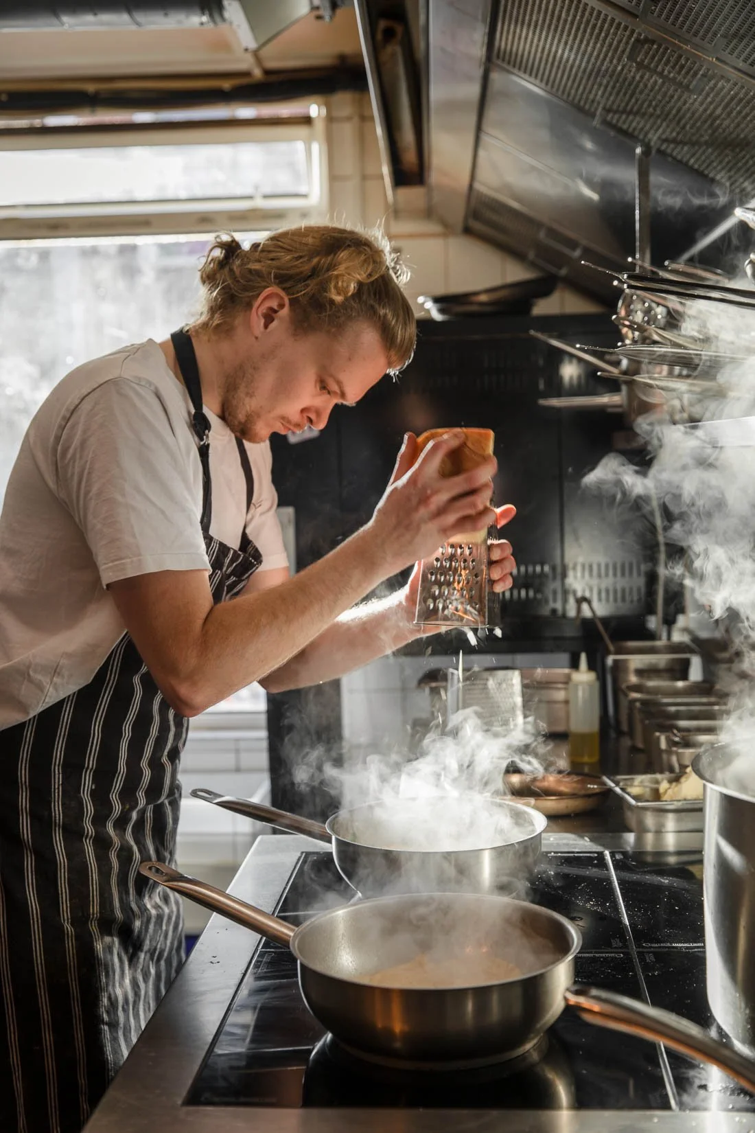 A chef adding grated cheese to a steaming pot in a professional kitchen.
