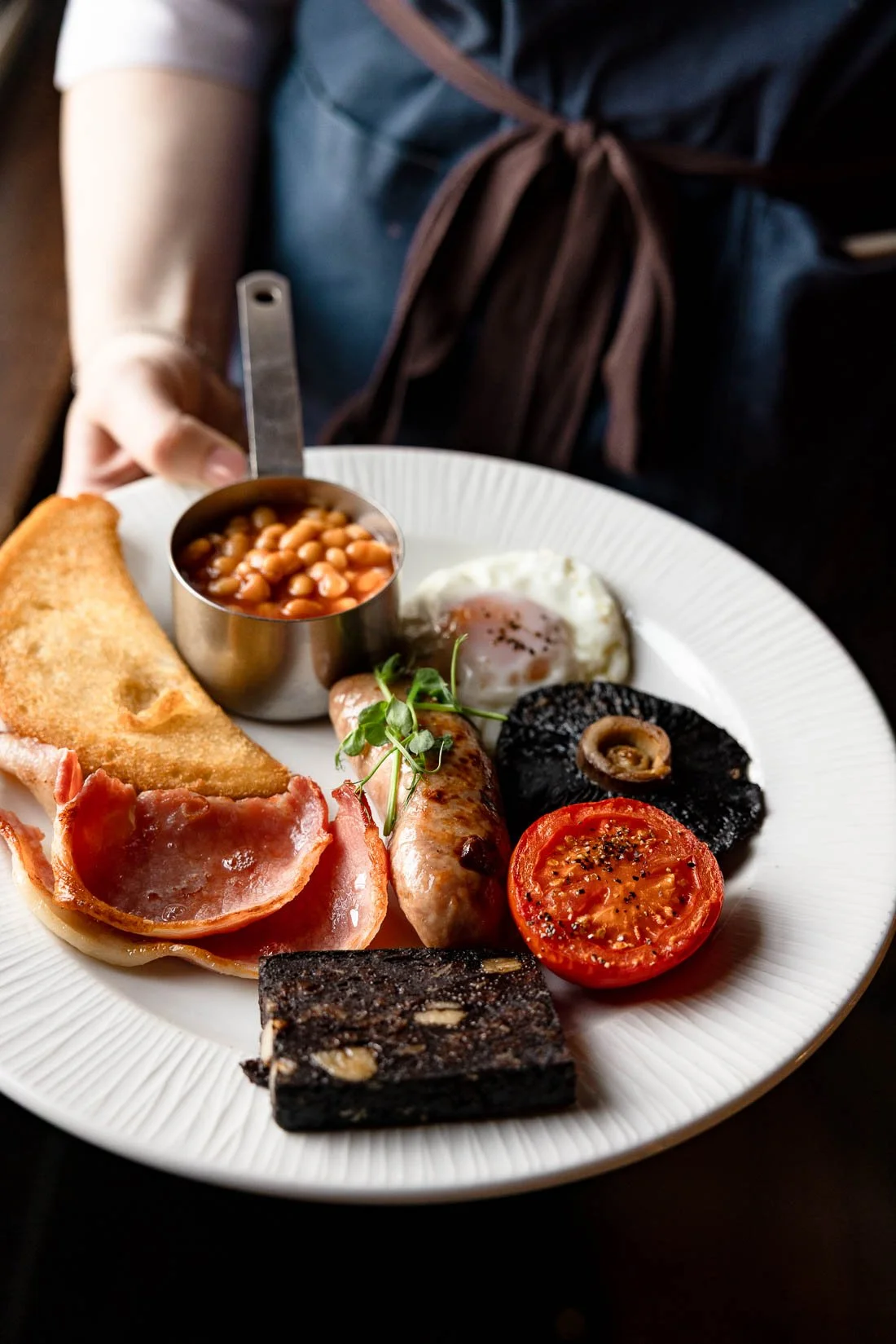 A white plate with a cooked breakfast including baked beans in a metal measuring cup, a fried egg, a sausage, a grilled tomato, black pudding, a slice of black pudding, crispy bacon, a buttery scone, and a small sprig of green herbs.