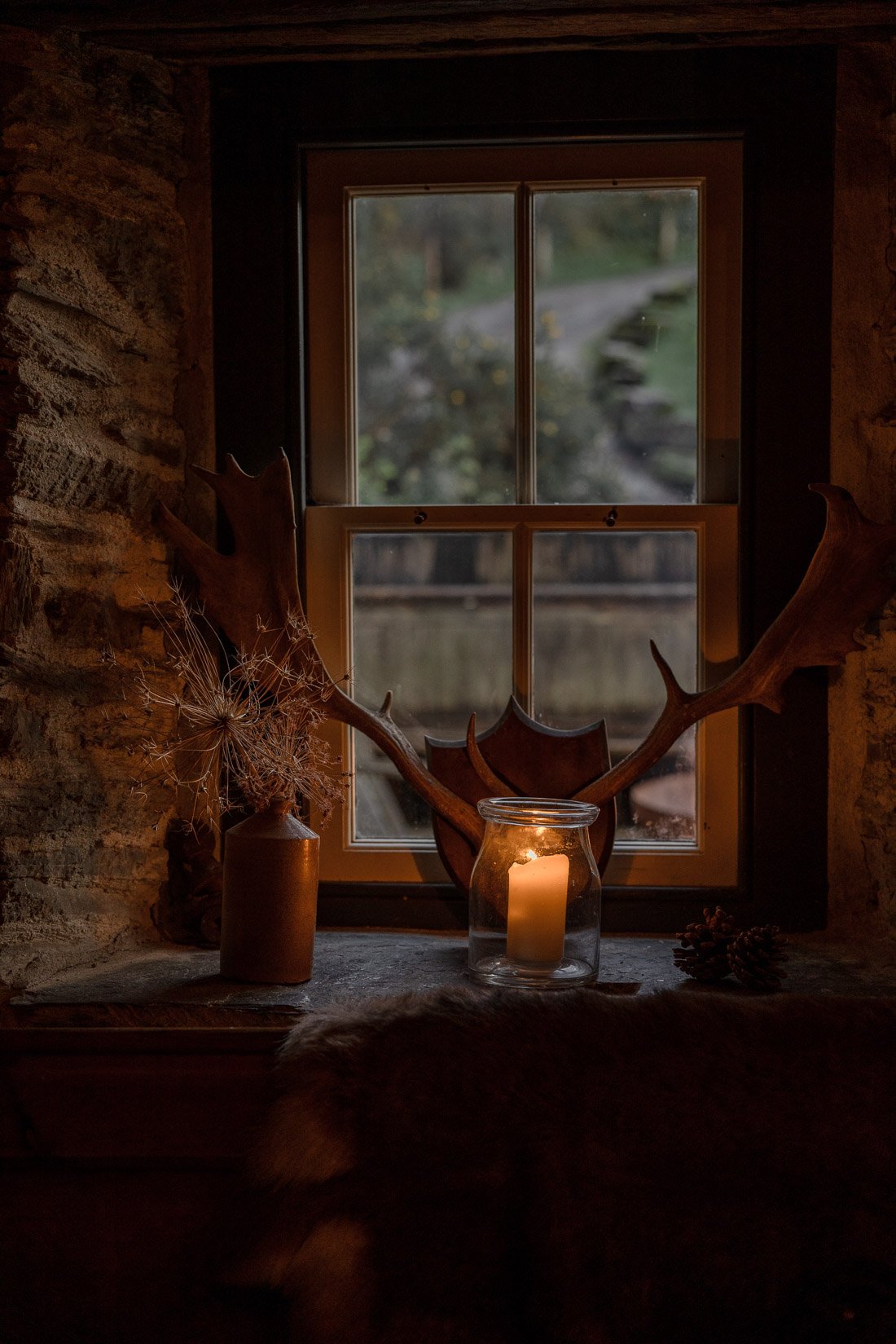 A cozy windowsill with antlers, a candle in a glass jar, a small pine cone, and a vase with dried plants, illuminated by a candle and looking out a window with a blurred outdoor scene.