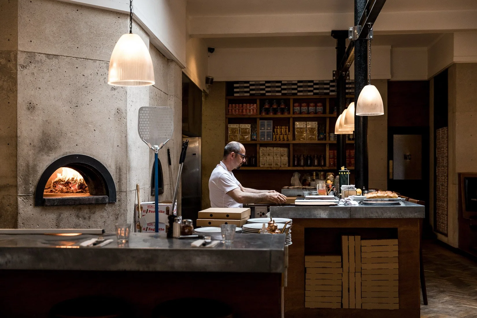 A man in a white shirt cooking at a kitchen counter in a cozy restaurant with warm lighting. There is an oven with a small flame on the left and shelves with jars and boxes in the background.