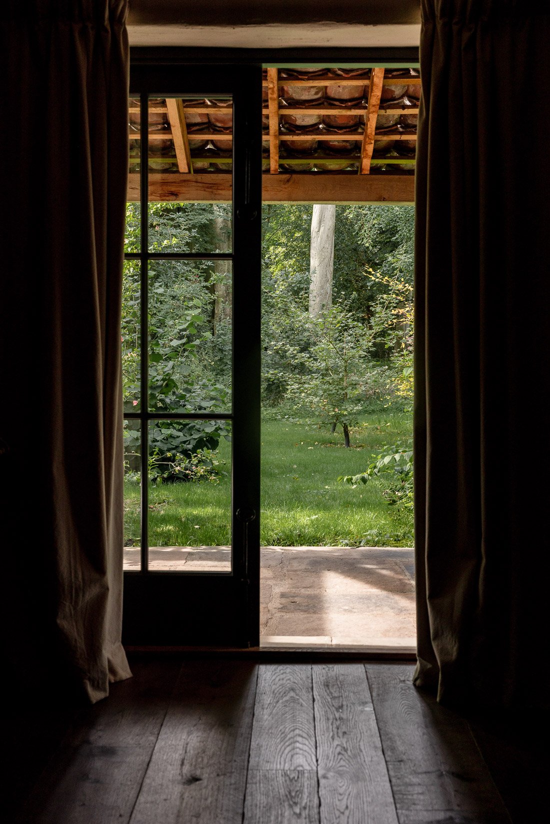 View through a glass door opening to a green backyard with trees and lawn, with curtains on either side.