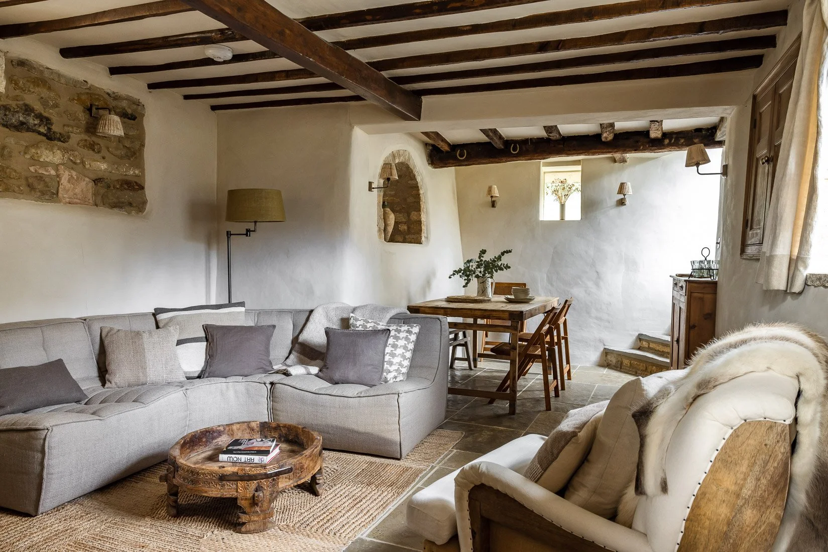 A cozy living room with a beige sofa, throw pillows, and a fur throw, a wooden dining table with chairs, stone walls, ceiling beams, a window with curtains, and decorative lighting fixtures.