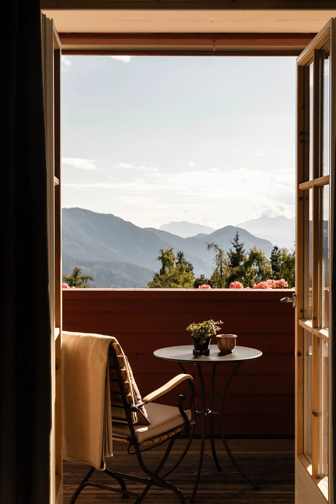 An open balcony door reveals a mountain view with trees and layers of distant mountains under a partly cloudy sky. The balcony has a small round table with a potted plant and a cup, a chair with a towel draped over it, and pink flowers on the railing