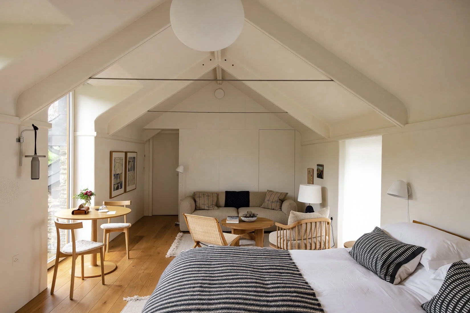 A cozy hotel room with a bed, a sitting area with a sofa and chairs, a round dining table with two chairs, and decor including framed artwork and lamps.