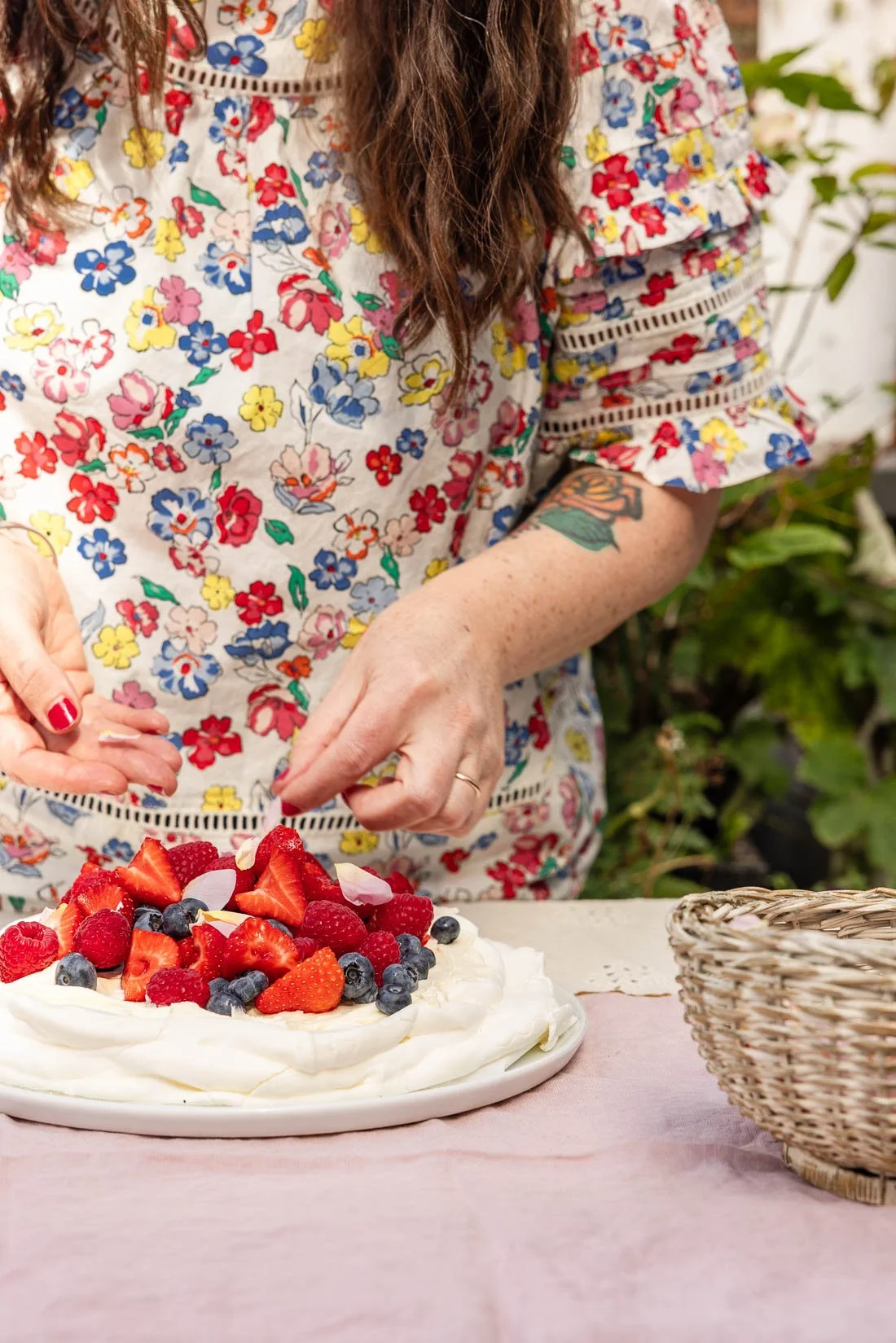 A woman in a colorful floral dress is arranging strawberries and berries on a white dessert topped with whipped cream, with a pink tablecloth and a wicker basket nearby.