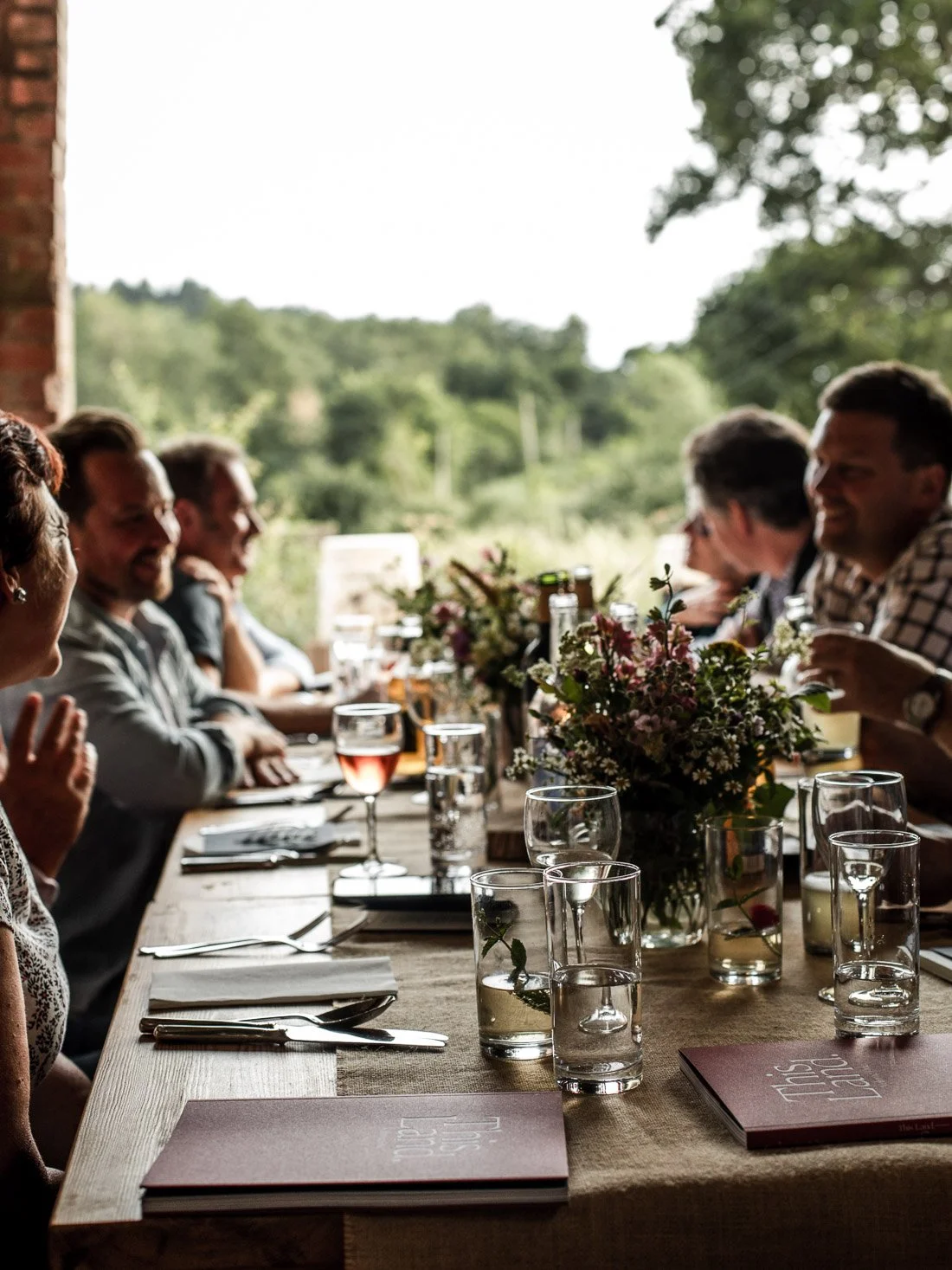 People sitting at a long wooden dining table with drinks and floral centerpiece, enjoying a gathering in a rustic setting with a view of green trees outside.