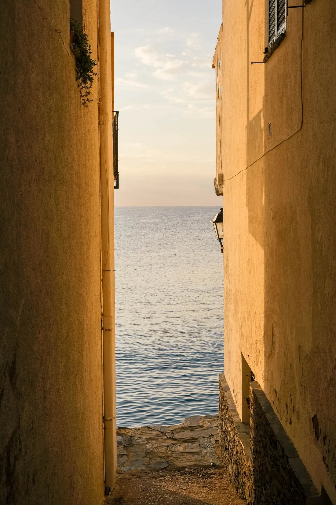 Narrow alleyway between two buildings with a view of the ocean or sea in the distance, and a partly cloudy sky above.