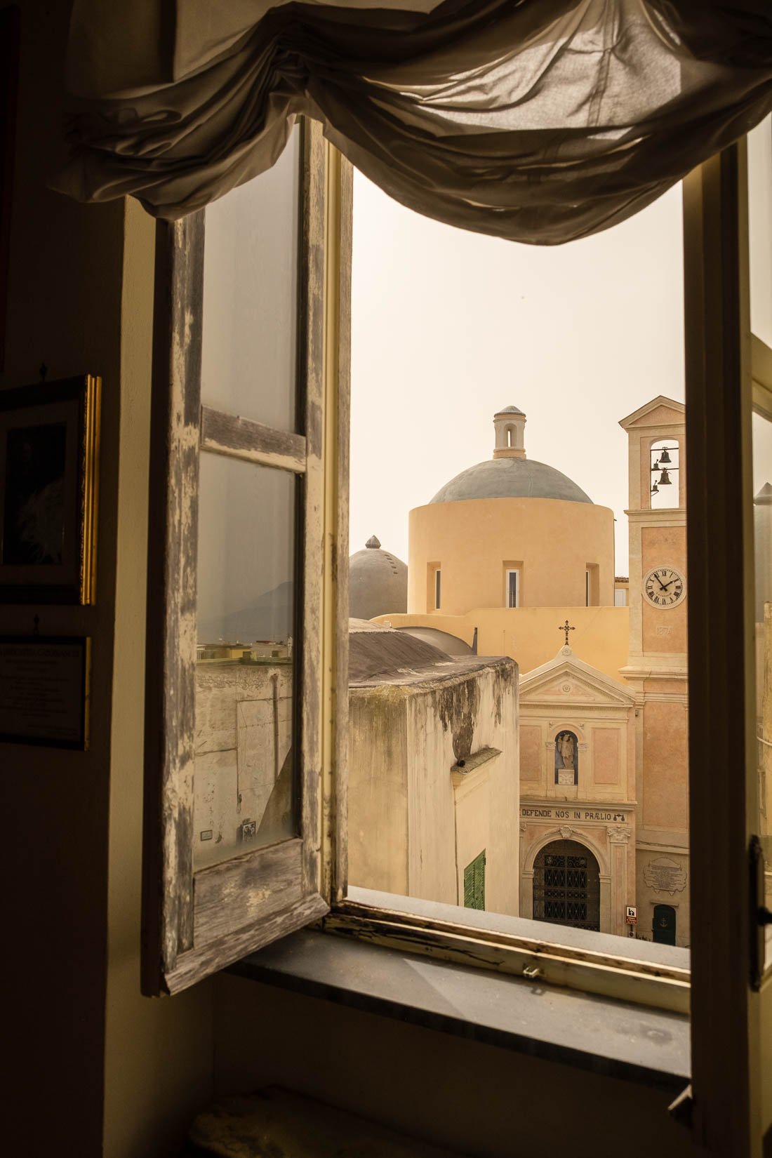 View of a historic European city through an open window, showcasing a church with a round dome, bell tower, and clock on a sunny day.