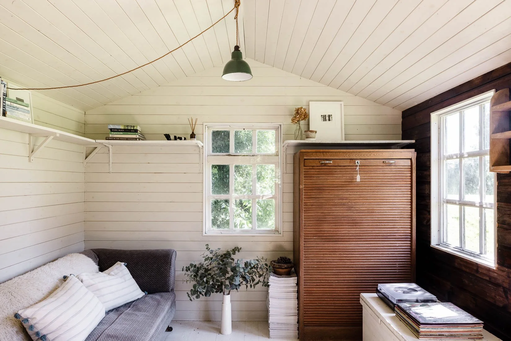 A cozy corner of a room with white wooden walls and a ceiling, a window with greenery outside, a gray armchair with white pillows, a potted plant, a white shelf with books, and a wooden roll-up cabinet.