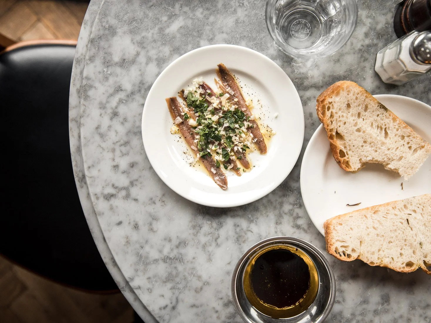 A top-down view of a marble table with a plate of anchovies garnished with chopped herbs and garlic, slices of bread on a plate, a glass of water, a bottle of olive oil, and a salt and pepper shaker.