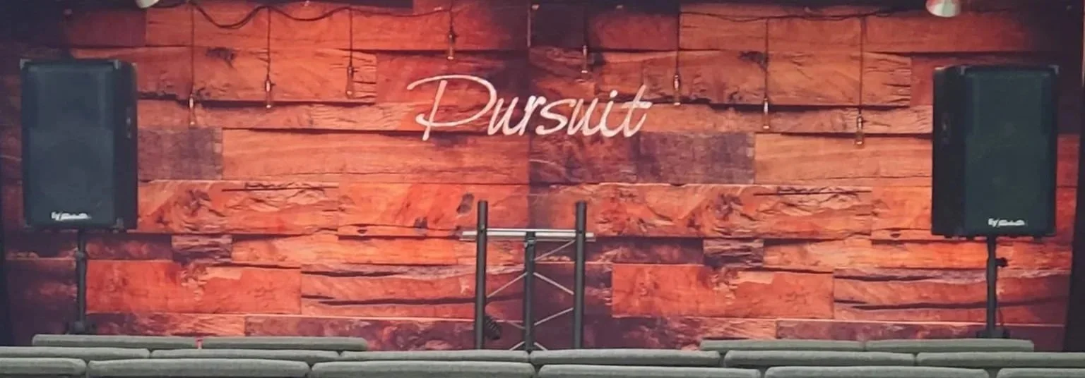 Stage with a wooden backdrop and a sign that reads 'Pursuit,' two large speakers on stands, and a metal podium in front.