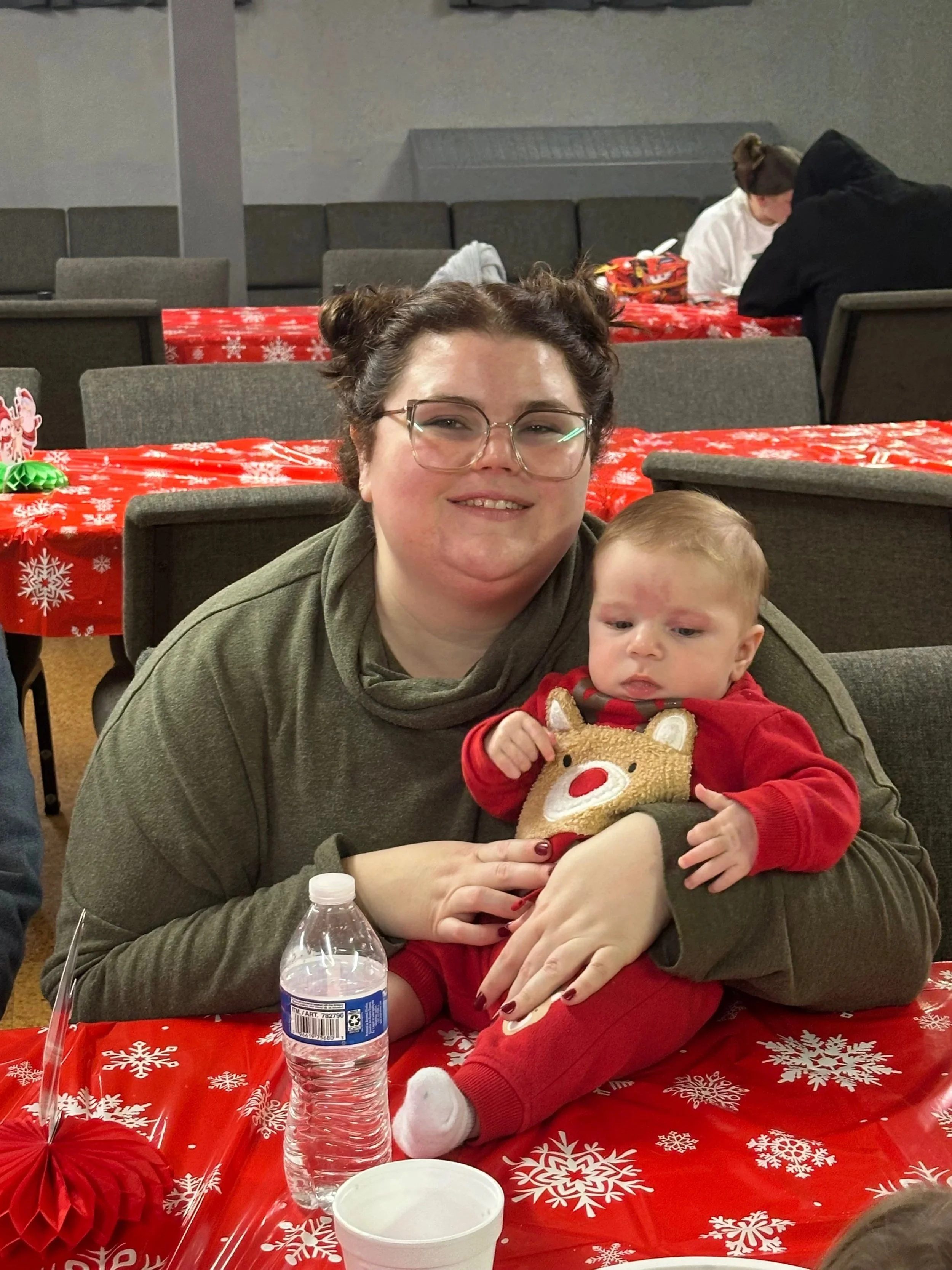 A woman with glasses and her hair in buns sitting at a table with a baby dressed in Christmas attire, surrounded by Christmas decorations.