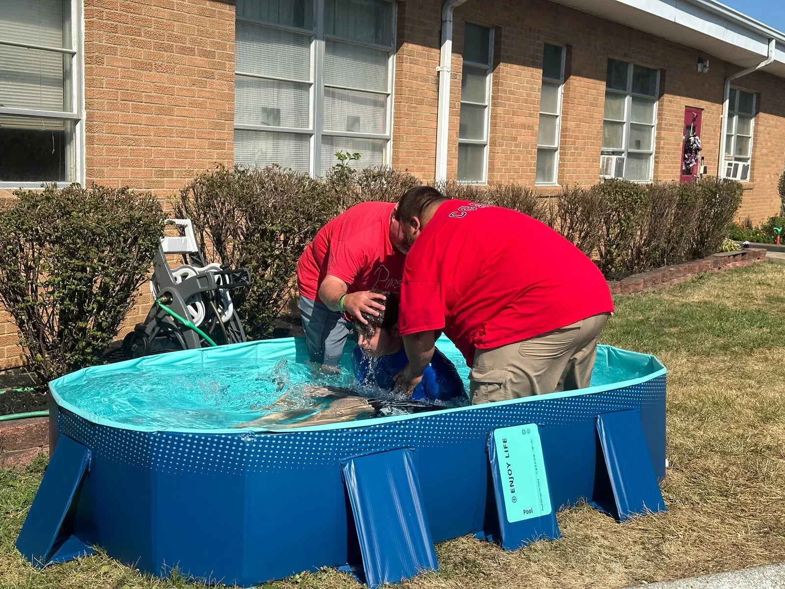 Two men in red shirts baptize a person in a small blue inflatable pool outside a brick building with bushes in the background.
